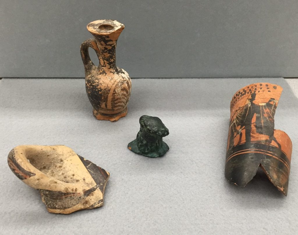 Object IDs (left to right) 1902.722.18: pottery sherd with handle attached; 2005.5.47: reconstructed lekythos vessel (circa 4th century BC); 2005.5.42: terracotta bust painted to look like patinaed bronze; 1902.722.4: reconstructed lekythos vessel. All objects were collected in Greece or other unknown parts of Europe in the mid- to late-1800s by Professor James Cooke Van Benschoten. All objects were donated by relatives of Professor Van Benschoten.